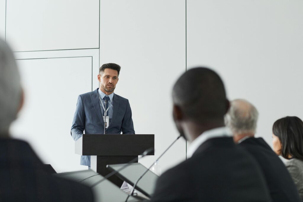 Speaker at a conference delivering a presentation to a diverse audience in a modern setting.